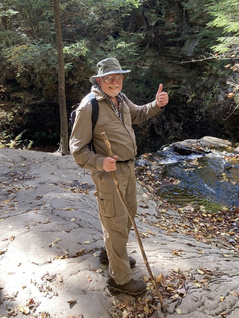 Bob Beck stands on a rocky hilside smiling, with a walking stick, sun hat, backpack and gives a thumbs-up. Photo by Gwen Beck.