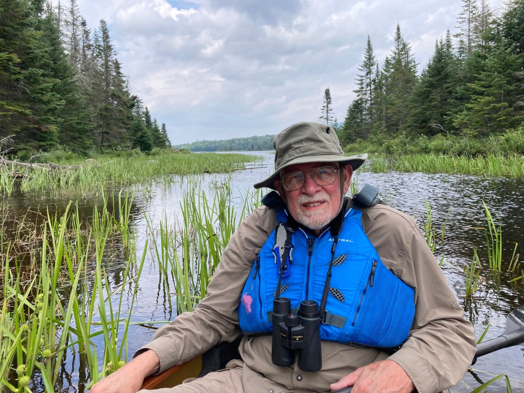 Bob Beck sits in a kayak on a river in the Adirondacks wearing binoculars, sun hat, and a life vest.